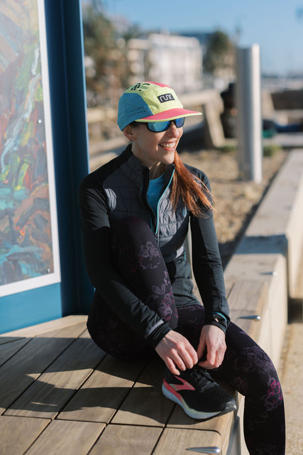 A woman wearing a colorful Runr Stuttgart Blackout Technical Running Cap, sitting on a wooden bench outdoors, smiling while dressed in sports attire with athletic shoes.