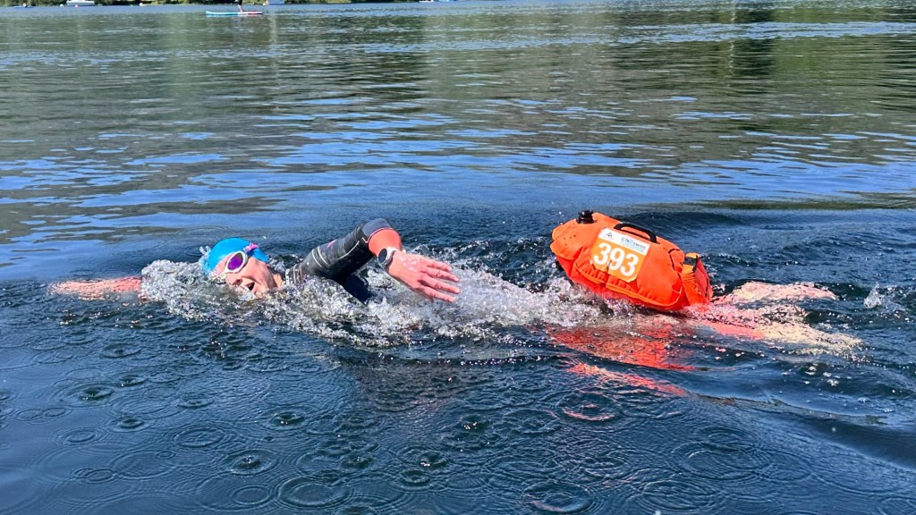 A swimmer wearing a bright blue swim cap and pink goggles swims in open water, with an orange buoy float attached to their waist.