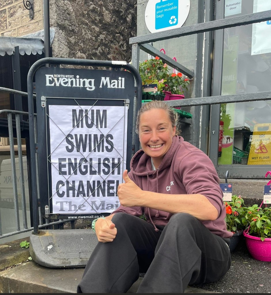 A smiling person giving a thumbs up while sitting next to a sign that reads 'MUM SWIMS ENGLISH CHANNEL', with potted plants in the background.