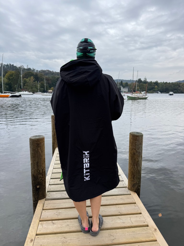 A Kitbrix all weather swimming changing robe  for women wearing a black KitBrix changing robe stands on a wooden dock by a lake, with boats in the background and a cloudy sky above.