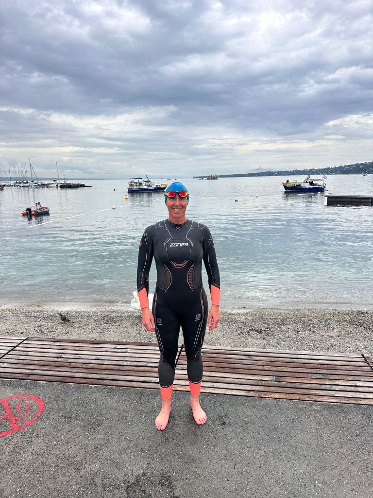 A swimmer wearing a Zone 3 Vanquish wetsuit, standing on a beach with a calm water background and cloudy skies.