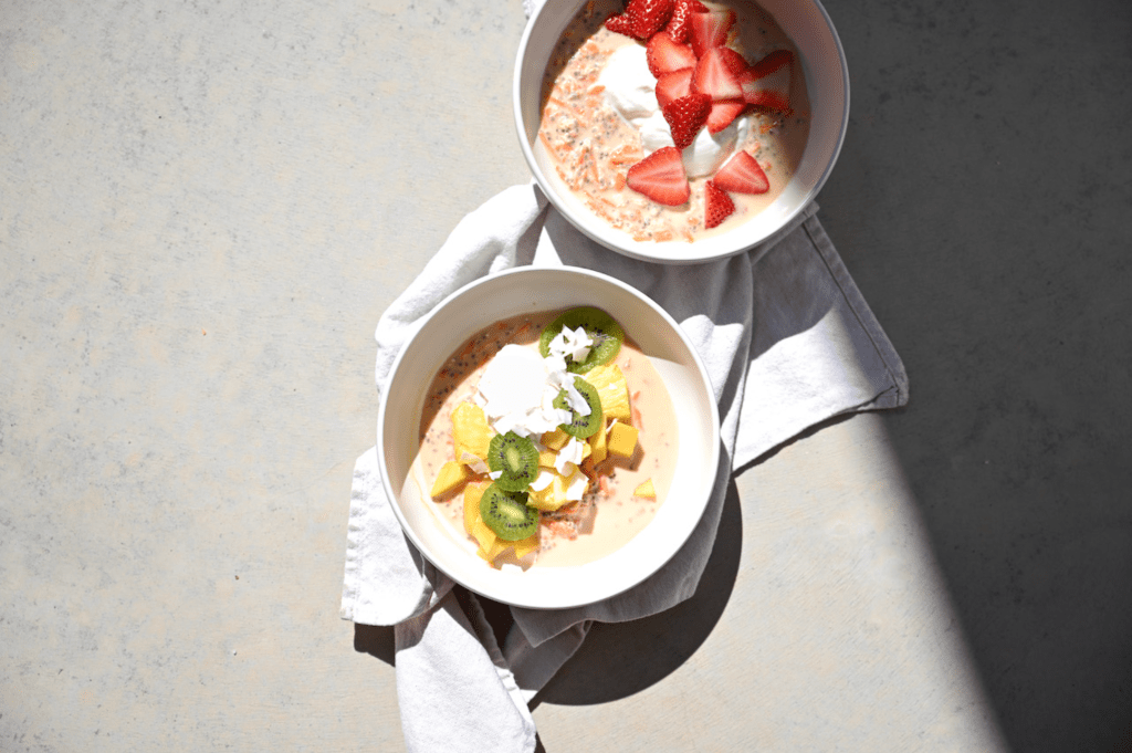 Two bowls of protein-rich smoothie bowls, one topped with strawberries and the other with kiwi and mango, placed on a light fabric.