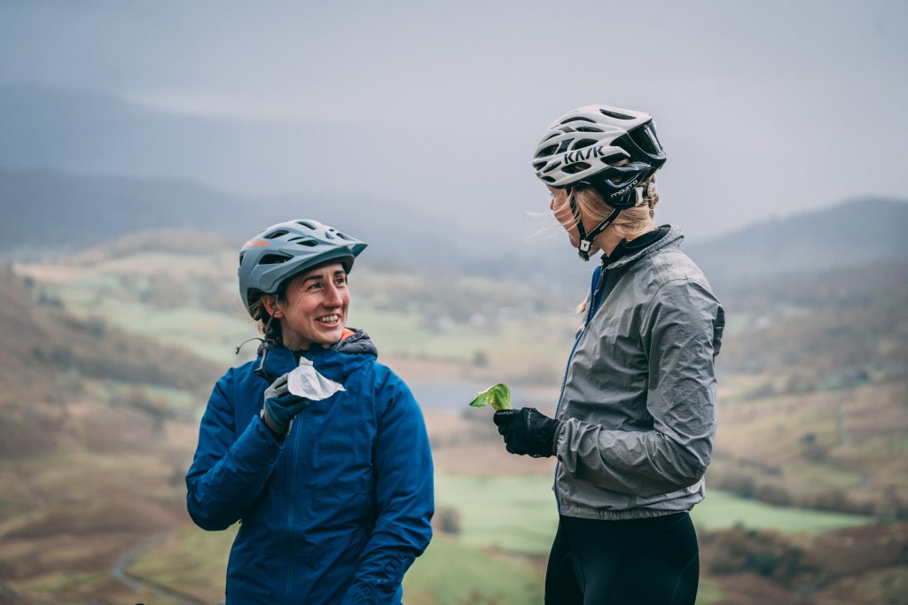 Two women wearing cycling helmets and jackets, standing outdoors with a scenic mountainous background, one holding an energy bar and the other holding a green snack.