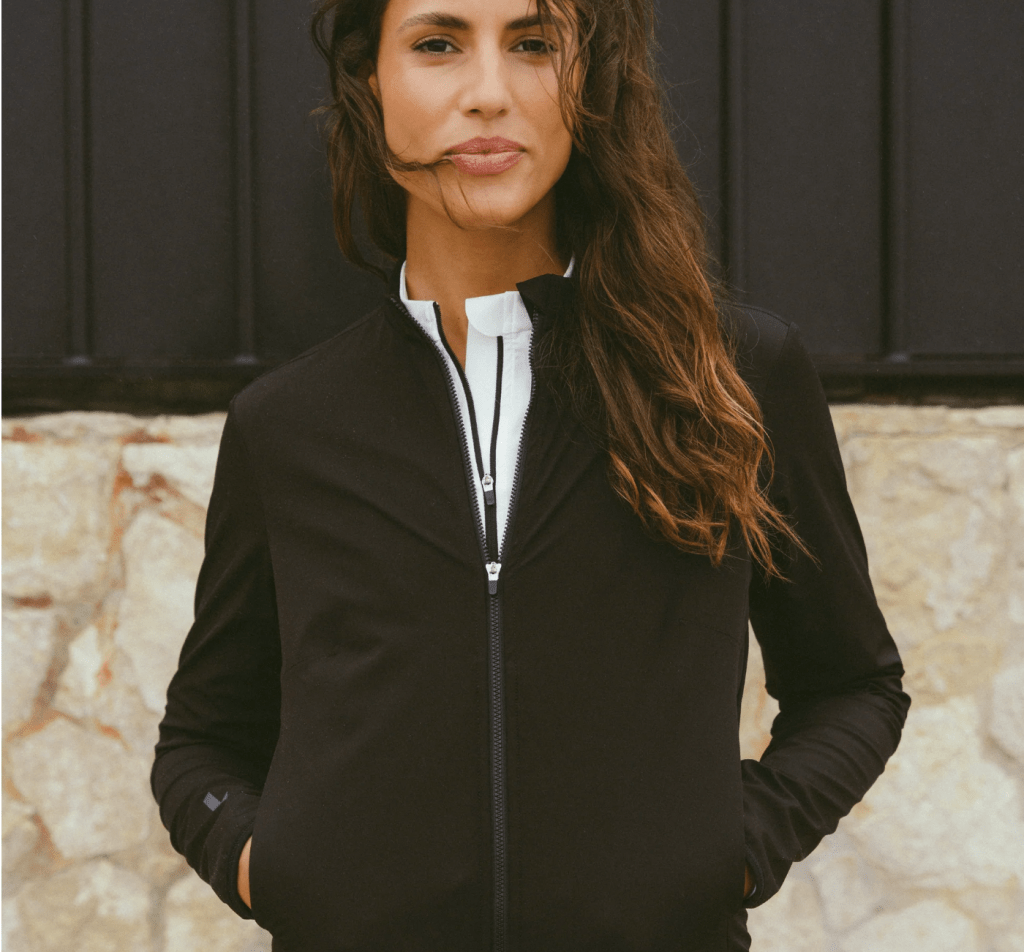 A woman wearing a fitted black cycling jacket with a zippered front, styled against a textured stone wall.