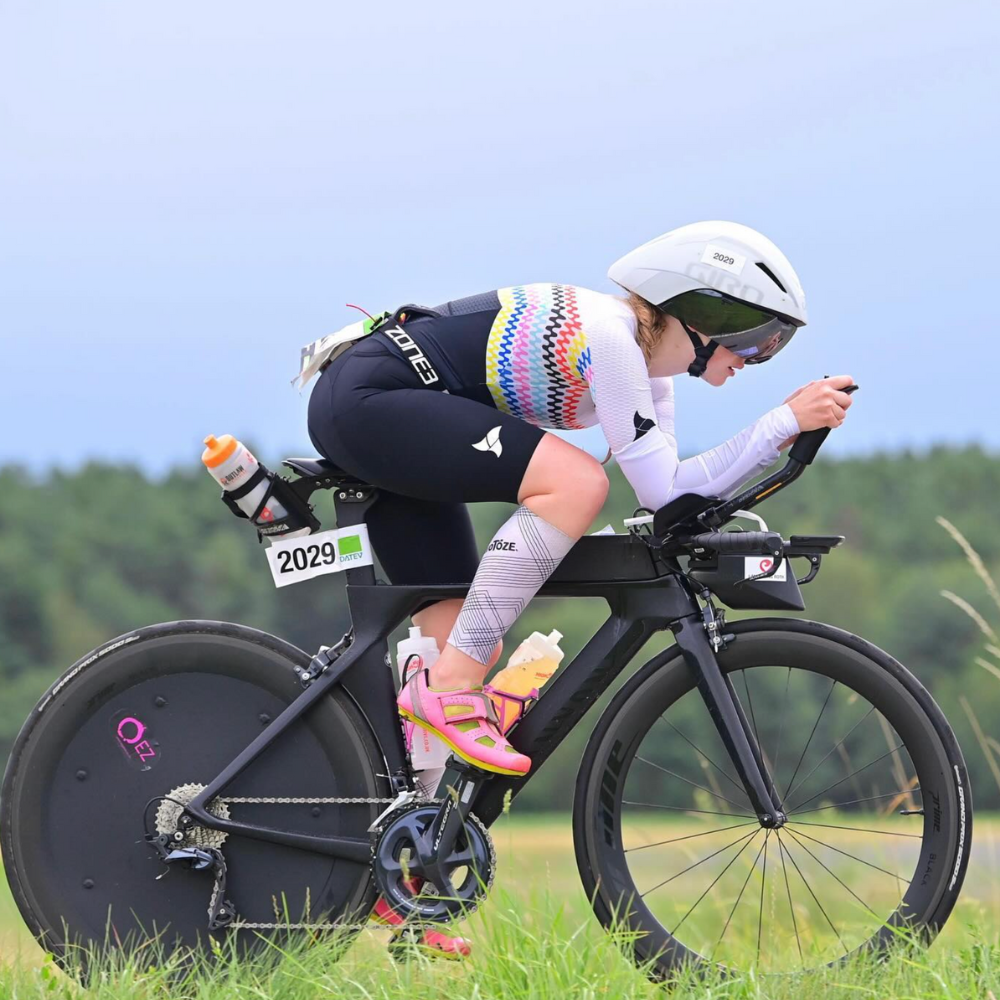 A female cyclist in a colorful jersey and helmet, positioned in a racing posture on a sleek black bike with a number 2029 displayed. The background features greenery and an overcast sky.