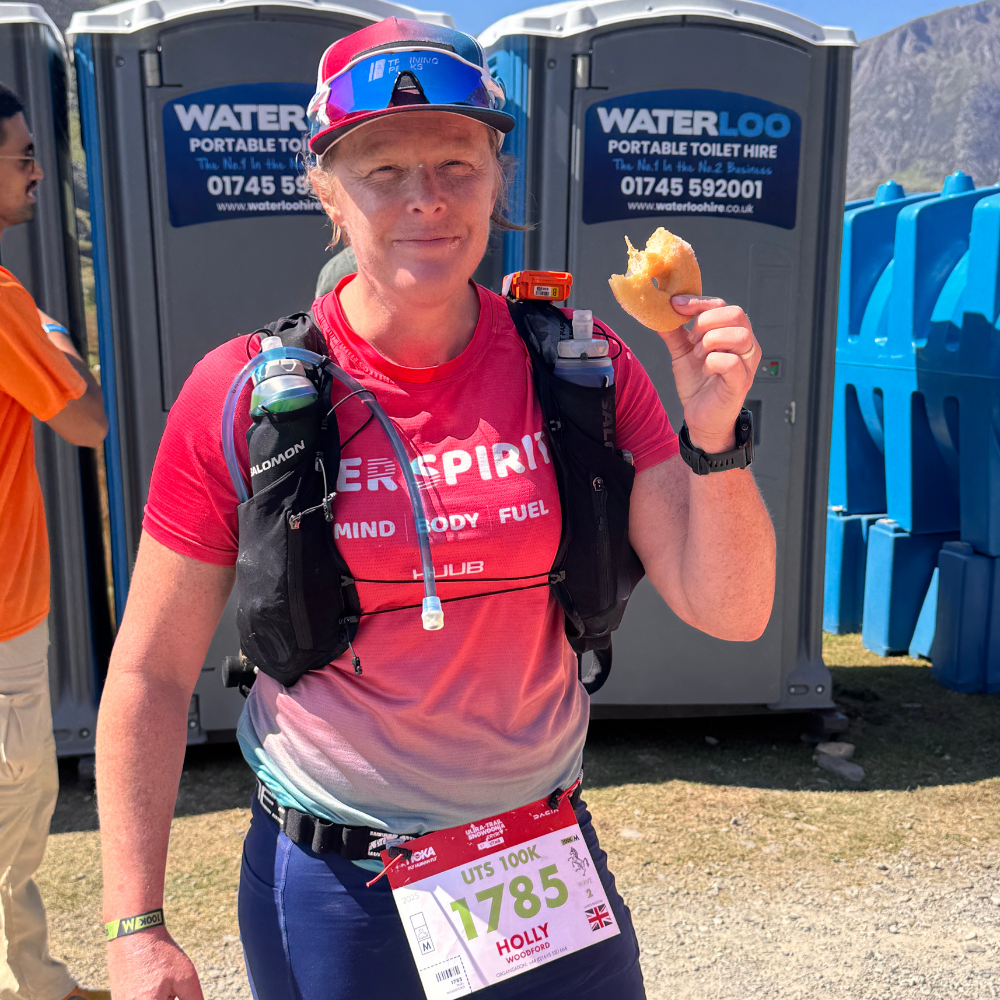 A woman wearing a running outfit and hydration pack, holding a piece of food, standing near portable toilets at an outdoor event.