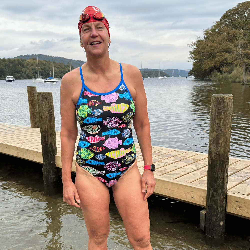 A woman smiling while standing on a dock near a lake, wearing a colorful fish-patterned swimsuit and swim cap.